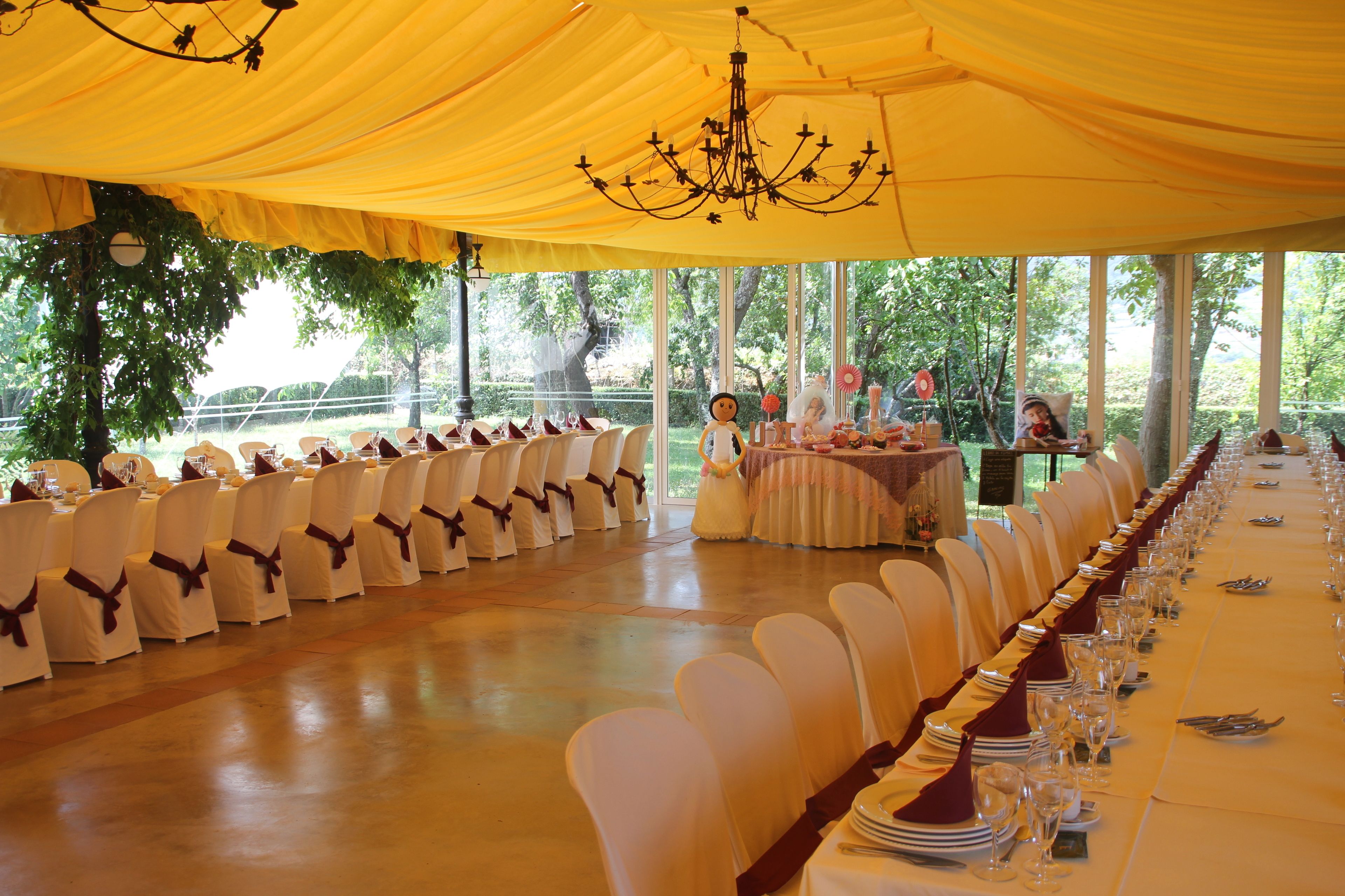 A banquet hall with white chairs and tables, a chandelier, and a view of trees outside.