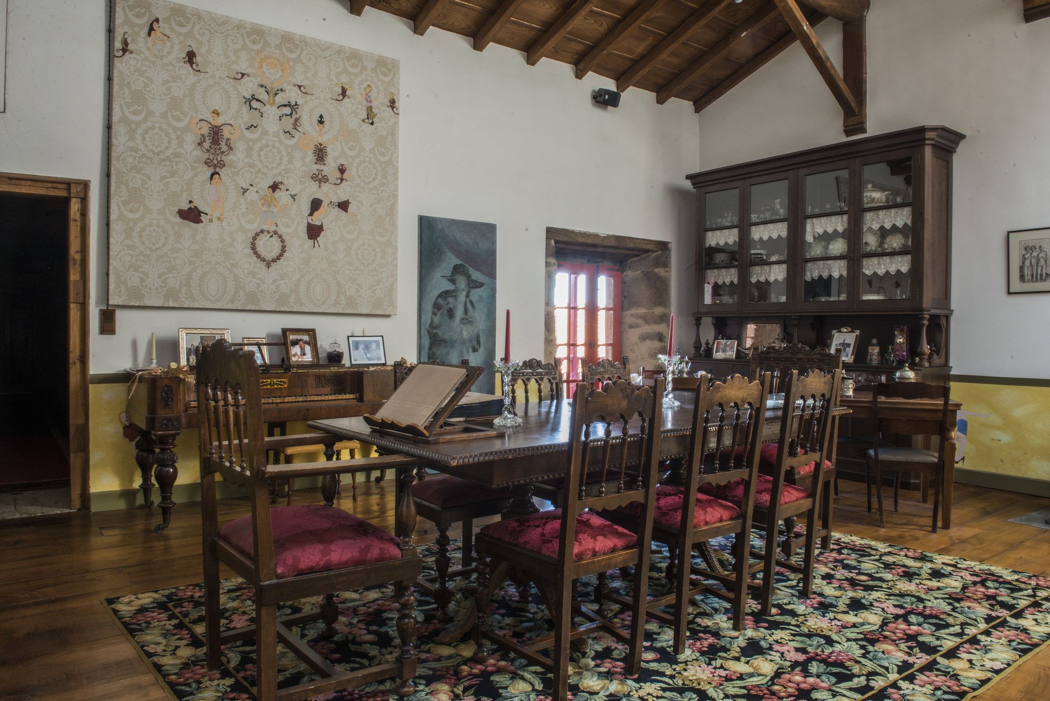 Un comedor con una mesa de madera, sillas tapizadas en rojo, un alfombra floral, un tapiz grande con motivos y un aparador de porcelana.