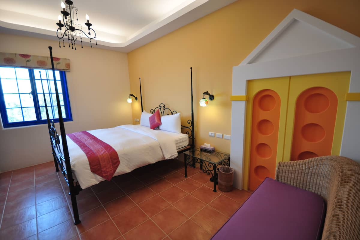 A bedroom with a bed, a nightstand, a chair, a window with blue curtains, and a wall-mounted light fixture.