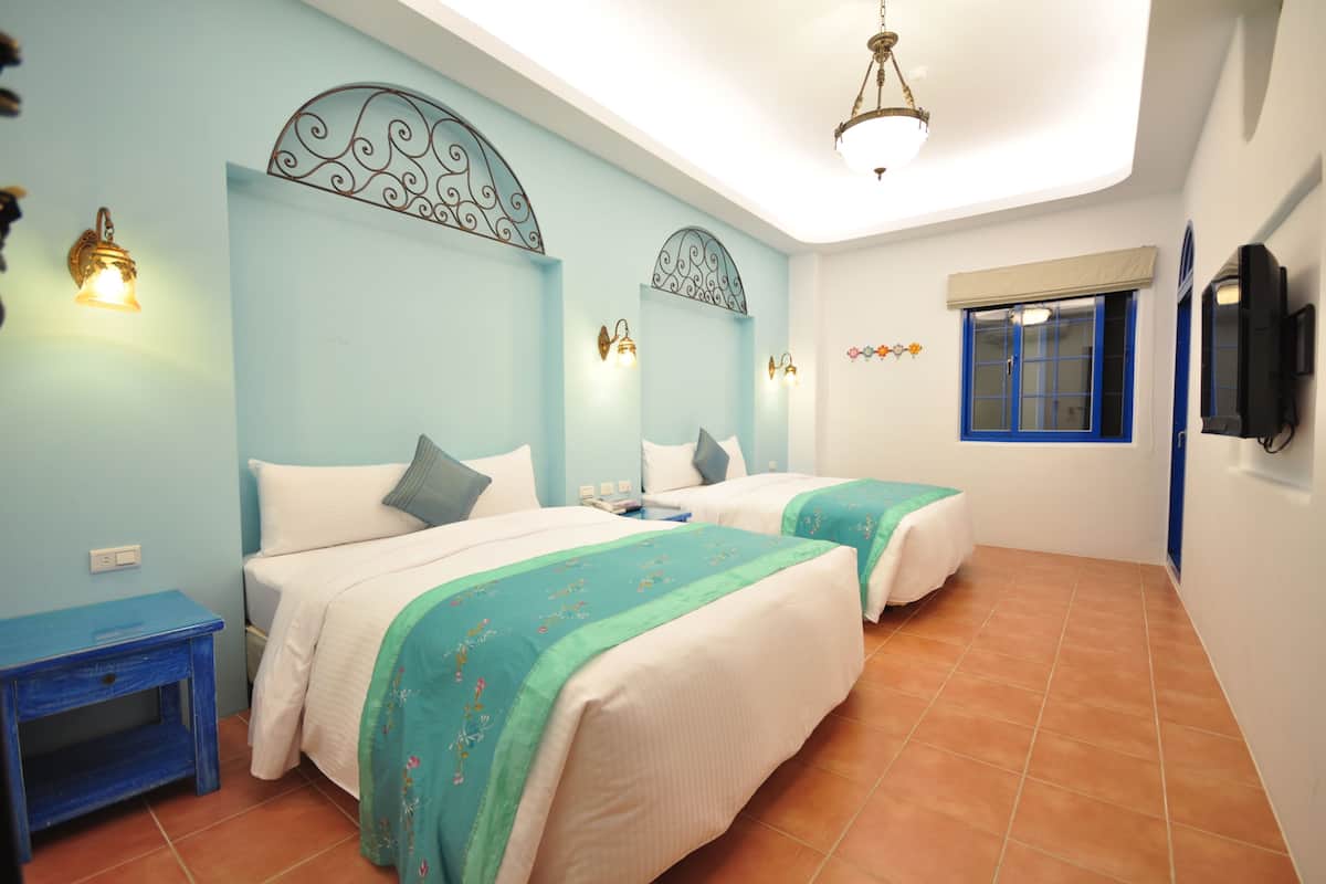 A bedroom with two beds, a small table, a window with blue shutters, and a ceiling light fixture.