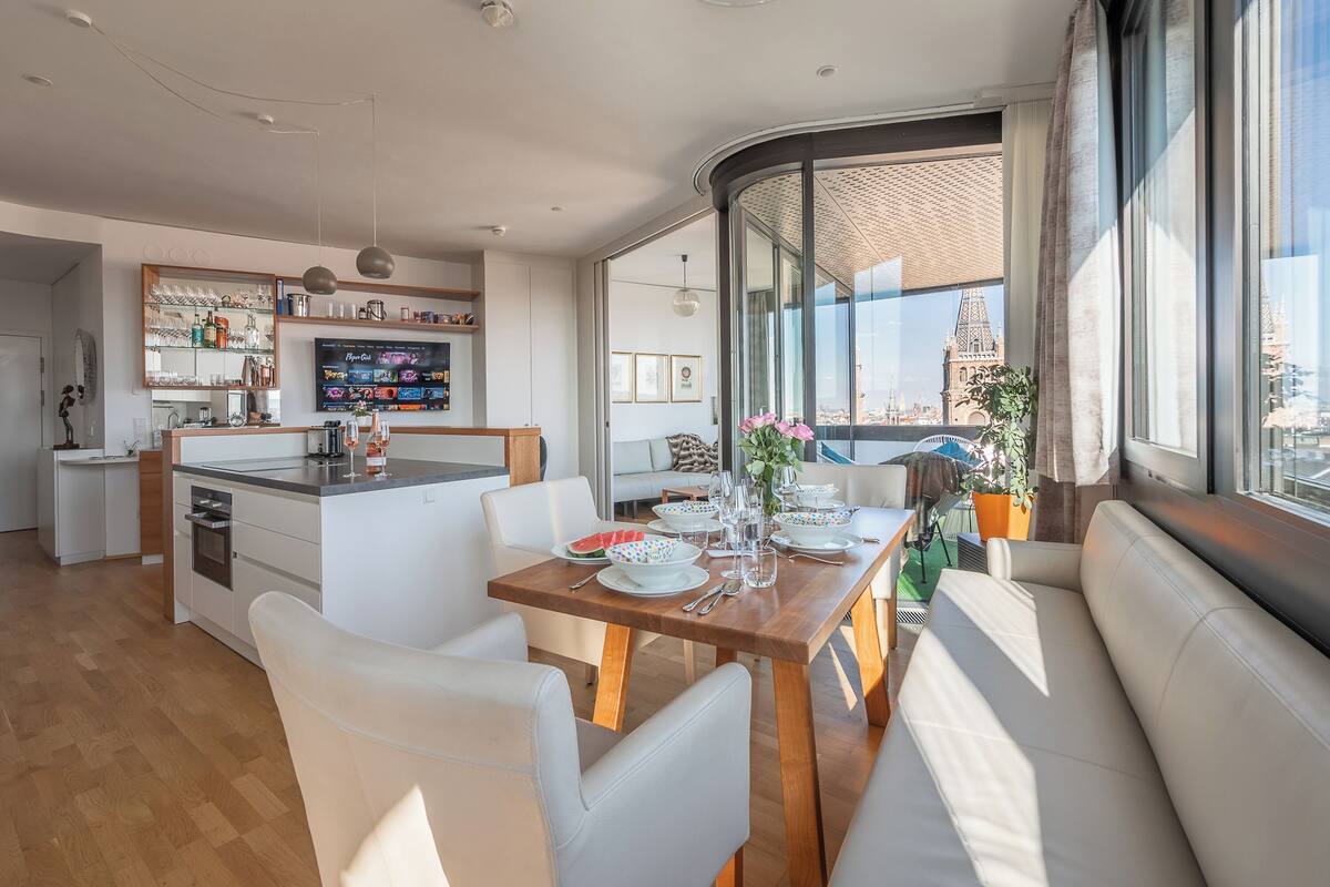 A modern kitchen with a dining area, large windows, and a view of a cityscape.