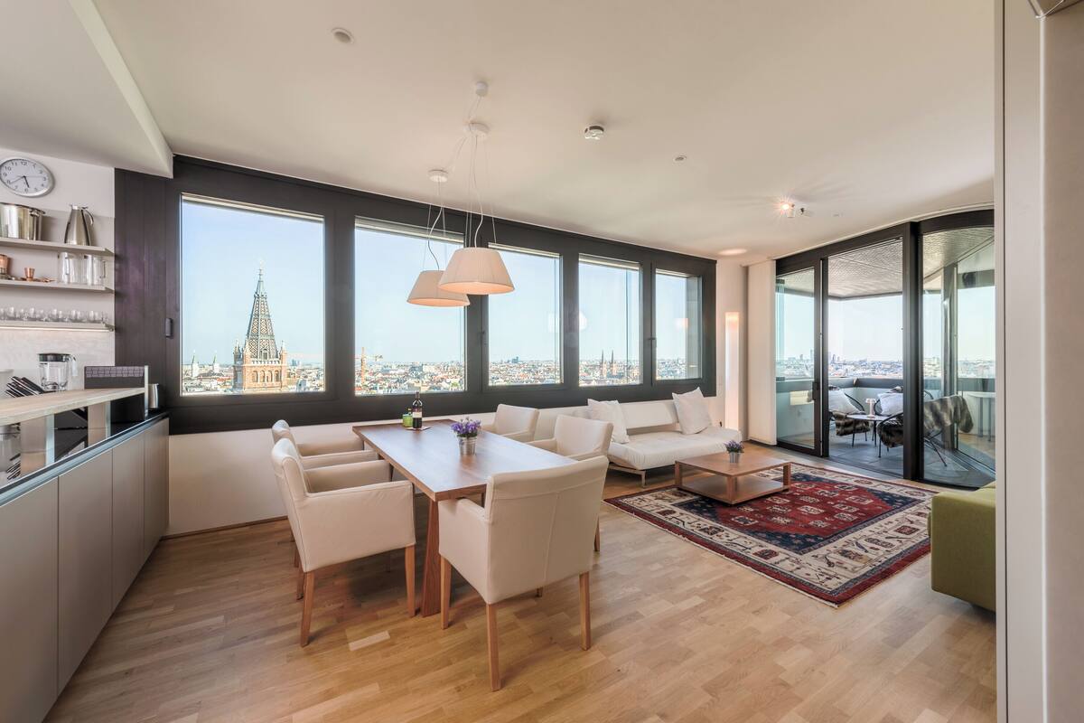 A modern living space with a dining area, sofa, and large windows offering a city view.