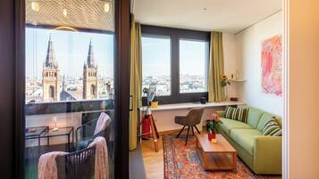 A modern living room with a green sofa, a wooden coffee table, and a view of a cityscape through large windows.