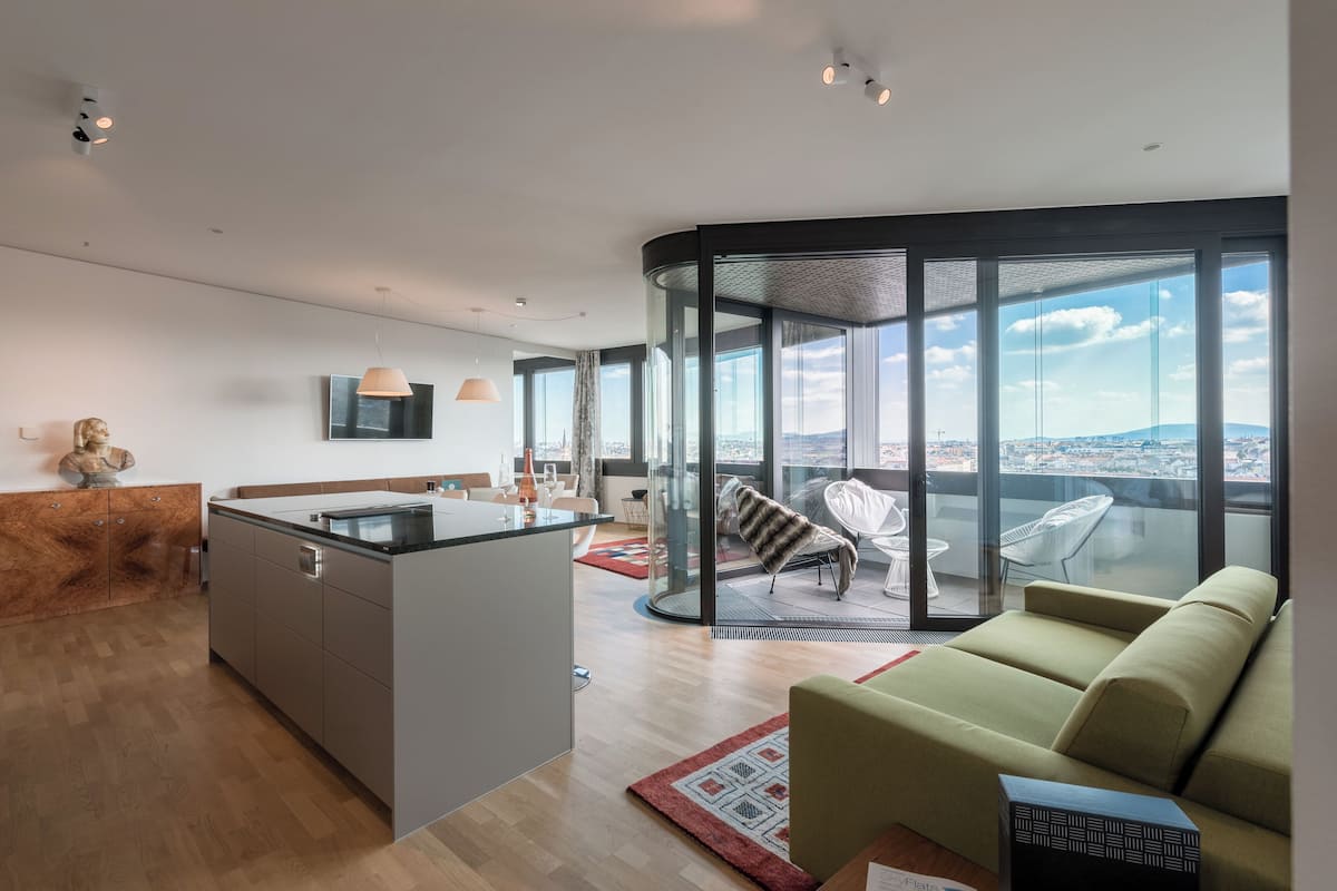 A modern living space with a kitchen, a seating area with a sofa and chairs, and a balcony with a city view.