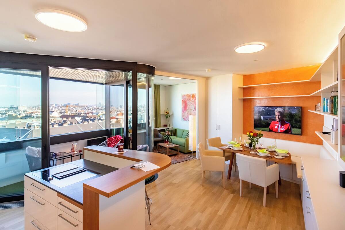 A modern hotel room with a large window, a dining area, and a TV screen displaying a sports event.