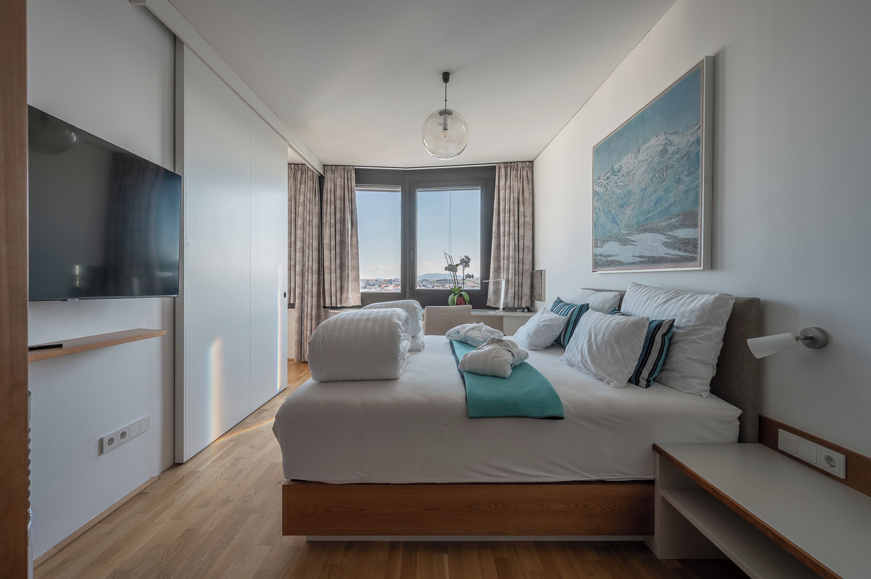 Una habitación de hotel moderna con una cama grande, un televisor, un cuadro y vistas a la ciudad a través de las ventanas.