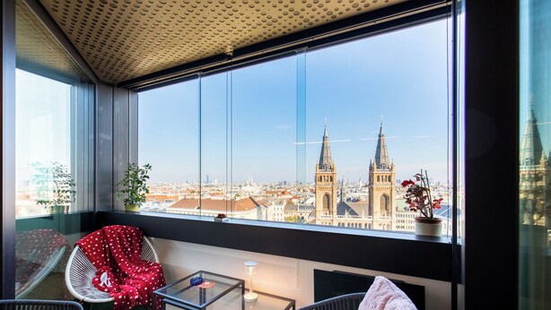 A balcony with a view of a cityscape, including two prominent towers, a potted plant, and a chair with a blanket. - Skyflats Vienna - Rooftop Apartments (Vienna)