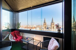 Un balcón con vistas a un paisaje urbano, incluyendo dos torres destacadas, una planta en maceta y una silla con una manta.