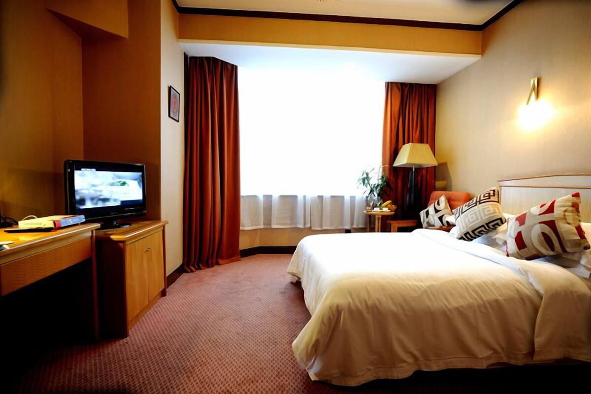 A hotel room with a bed, a desk with a television, a lamp, and a window with curtains.