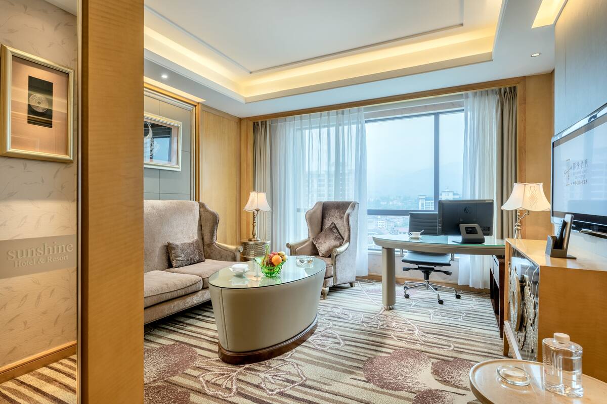 Ett hotellrum med en soffa, fåtölj, skrivbord, dator och ett stort fönster med gardiner.