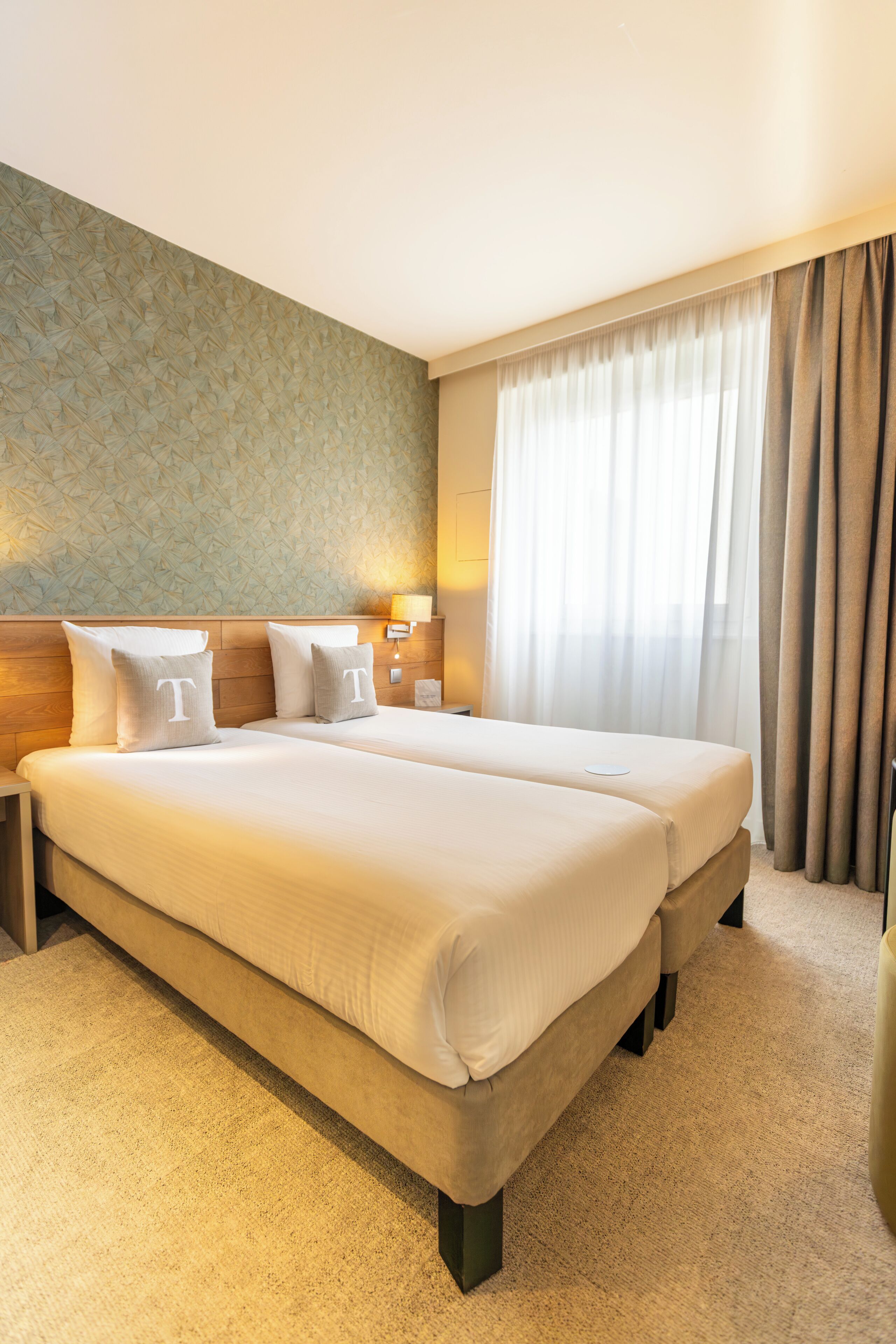 Ein Hotelzimmer mit zwei Betten, einer Wand mit Muster, einem Fenster mit Vorhängen und einem Nachttisch mit Lampe.