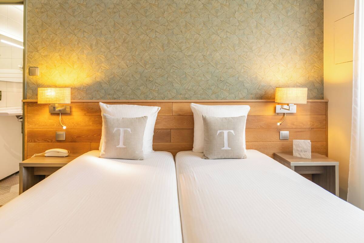 A hotel room with two beds, a wooden headboard, two bedside lamps, a phone on the nightstand, and a wall with a geometric pattern.