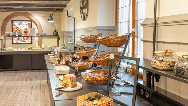 A buffet setup with various food items, baskets, and a clock on the wall. - Hôtel Maison Turenne (Colmar)