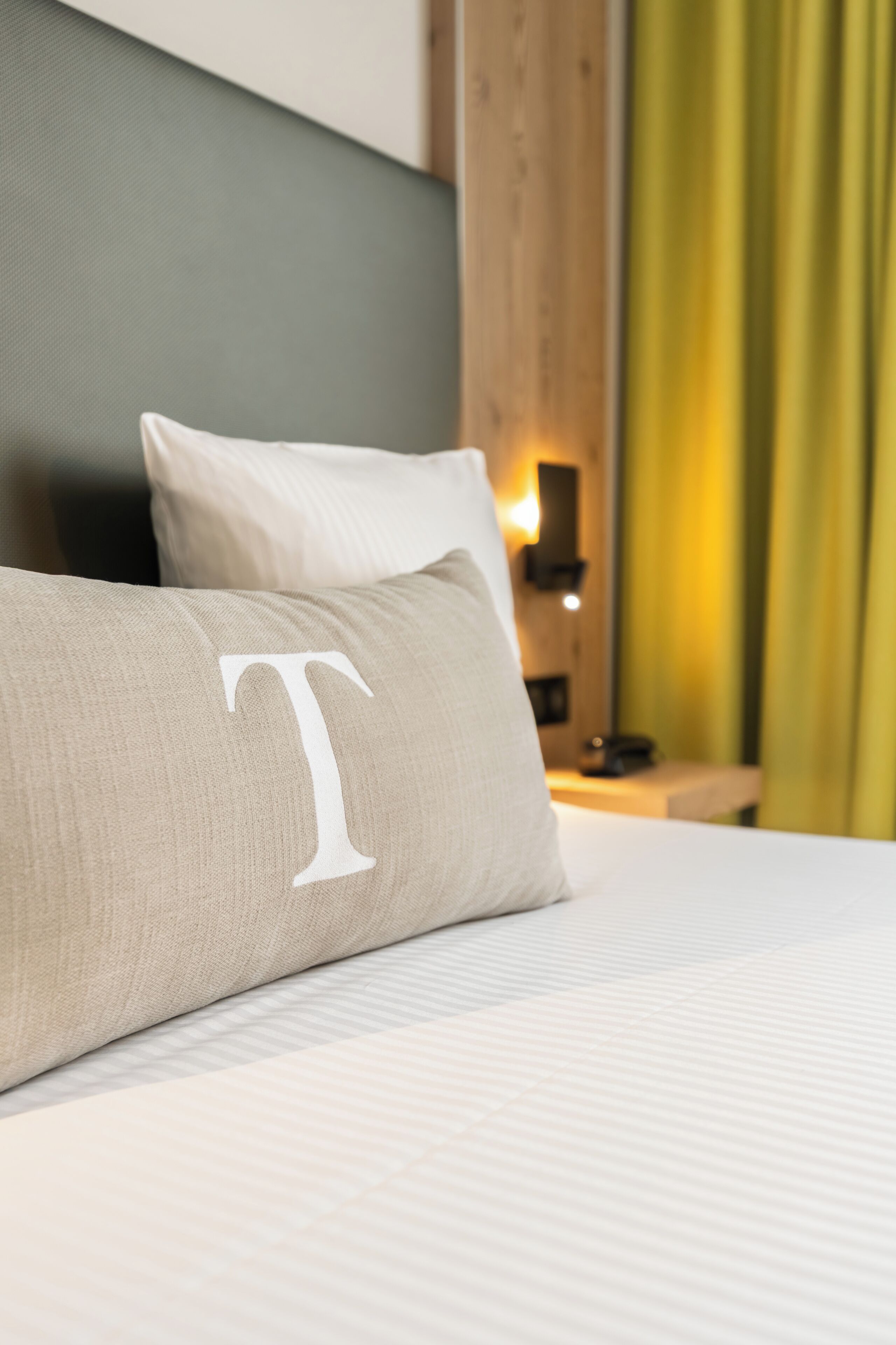 Ein Bett mit einem Kissen, das einen großen Buchstaben 'T' zeigt, und ein Nachttisch mit einer Lampe.