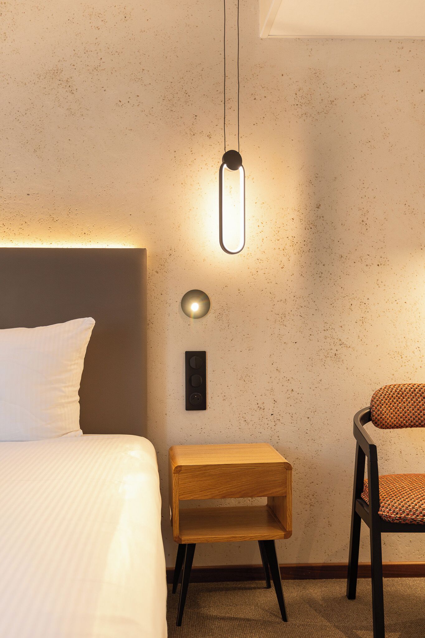 A neatly arranged hotel room with a bed, a small wooden side table, a chair, and a wall-mounted light fixture.
