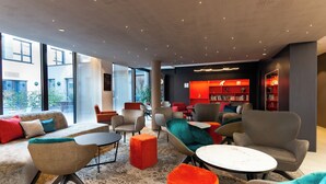 A modern lounge area with sofas, armchairs, and a coffee table, featuring a large window and a bookshelf with books. - Hôtel Maison Turenne (Colmar)