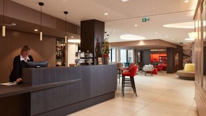 A reception area with a counter, a receptionist, and a seating area with red chairs. - Hôtel Maison Turenne (Colmar)
