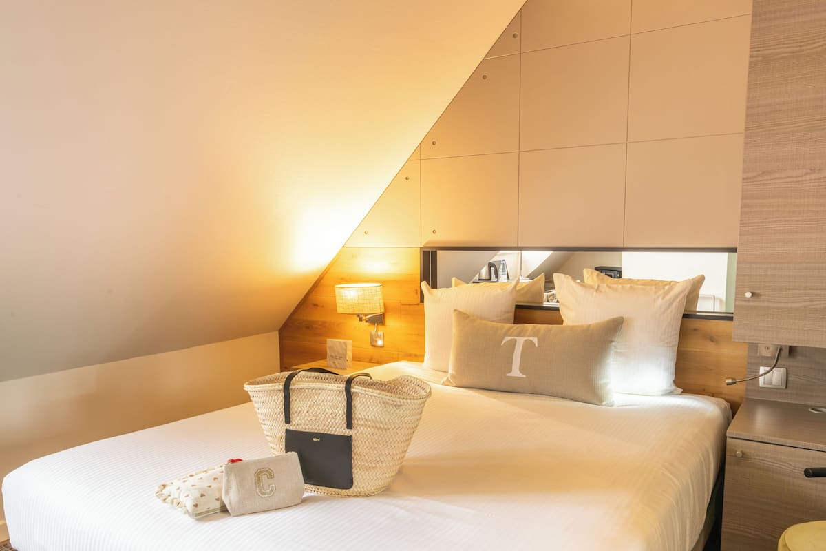 A hotel room with a bed, pillows, a nightstand, a lamp, a mirror, and a wooden headboard.