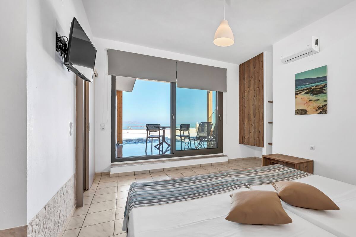 A modern hotel room with a large bed, a view of the beach, a TV mounted on the wall, and a wooden door.