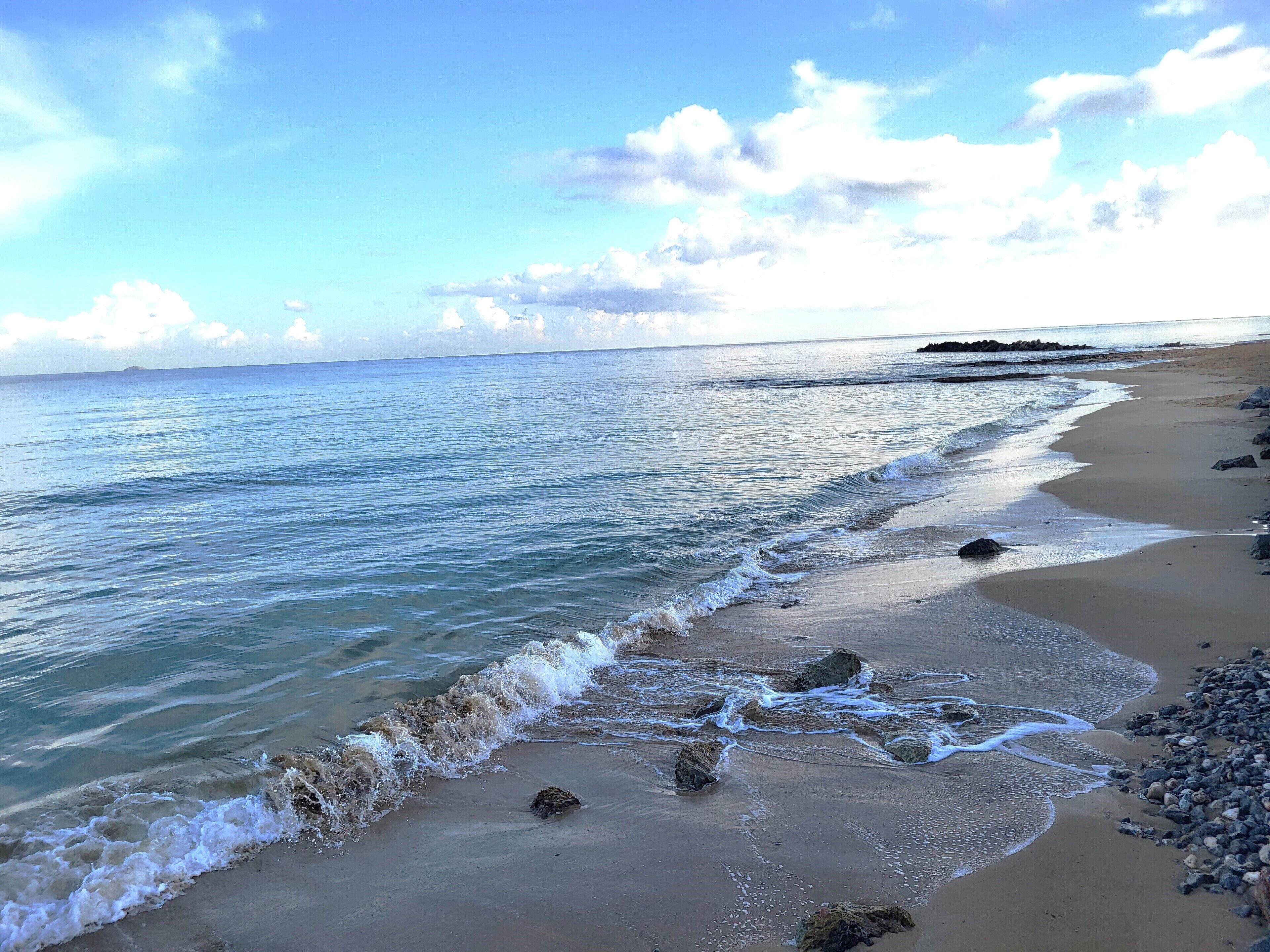Uma praia com águas azul-claras, areal arenoso e pedras espalhadas.