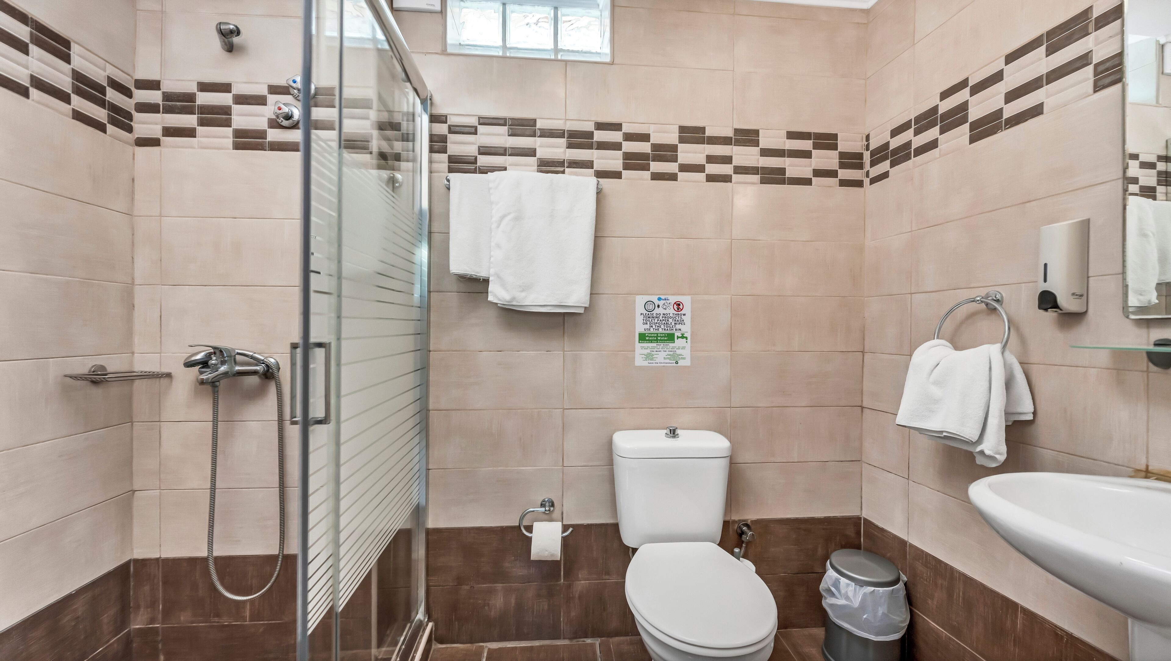 A bathroom with a glass shower enclosure, a toilet, a sink, and a trash bin.
