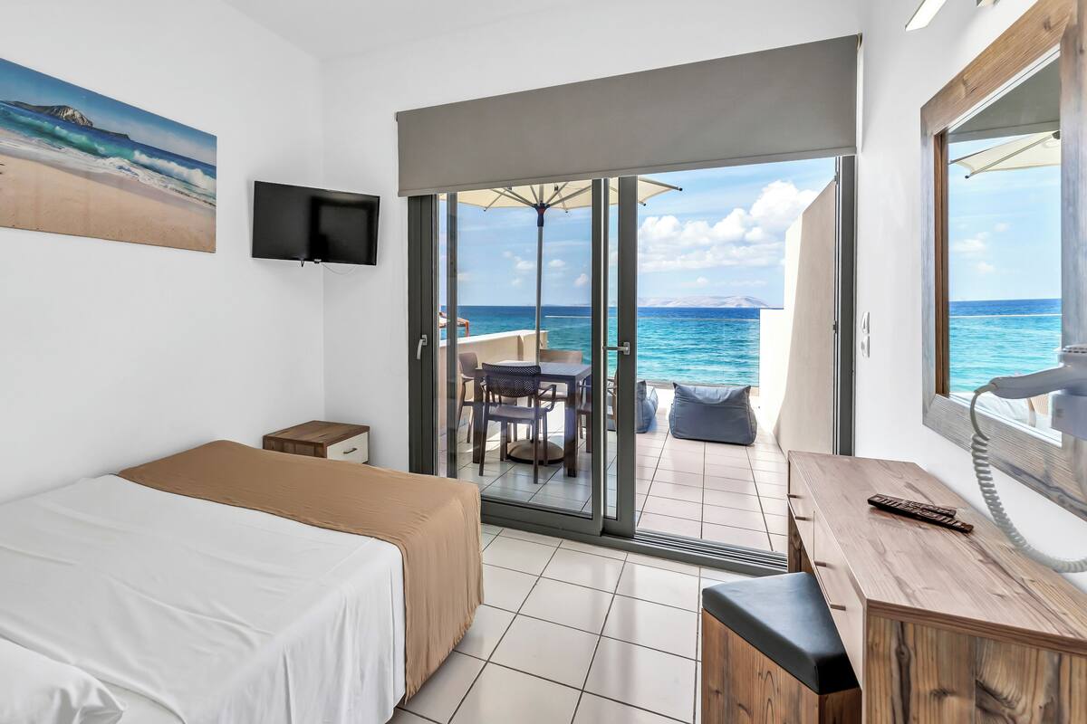 A hotel room with a bed, a desk, a chair, and a view of the ocean through a sliding glass door.
