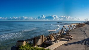 A coastal walkway with wooden benches and tables, a concrete path, and a view of the sea and sky. - Almare Beach Hotel (Hersonissos)