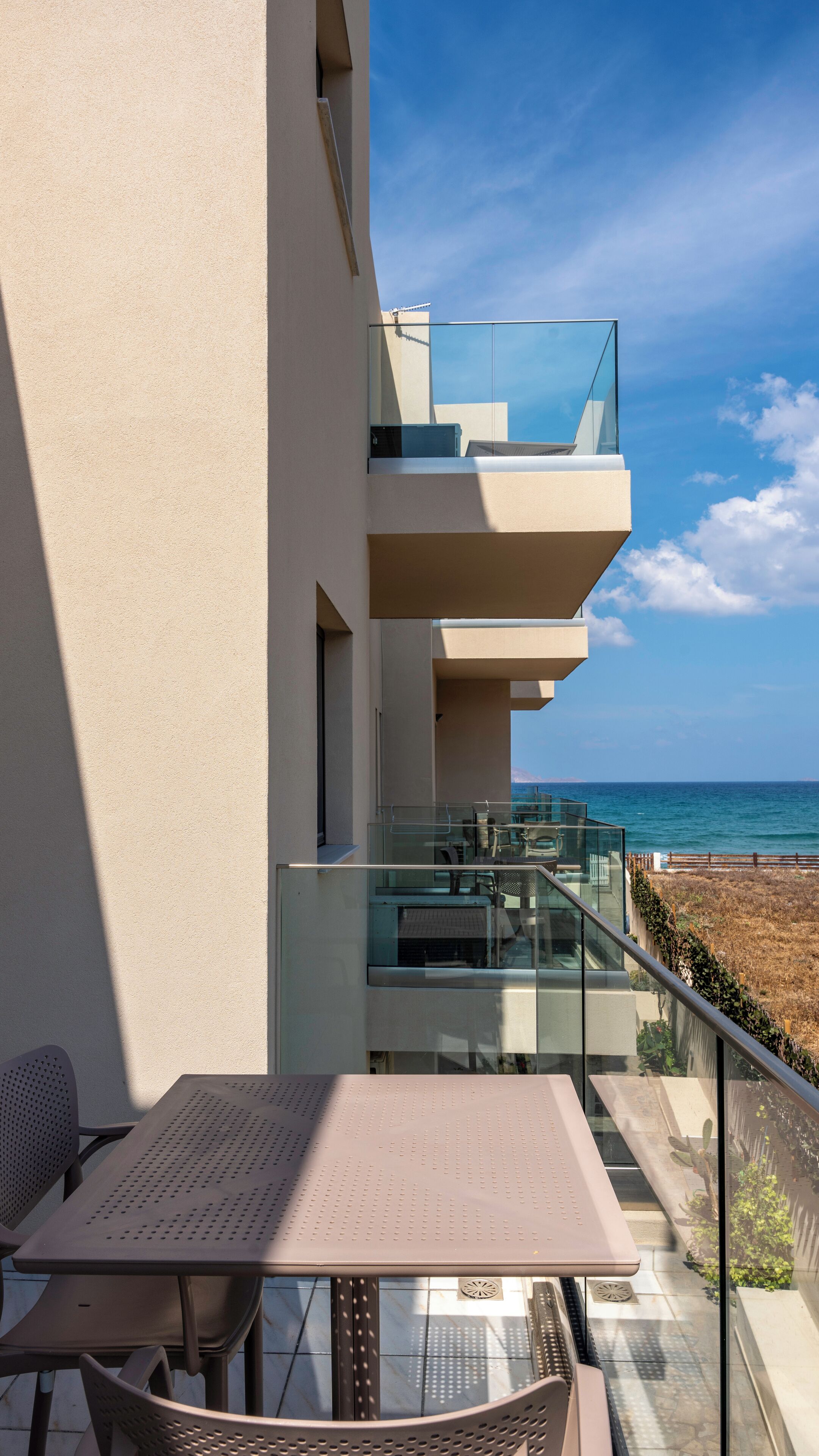 Un balcón moderno al aire libre con barandilla de vidrio, una mesa y sillas, con vistas a una playa y al océano.