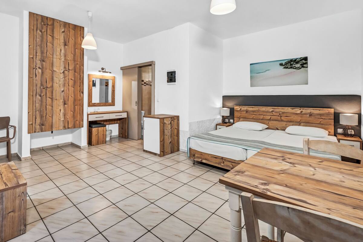 A modern hotel room with a wooden headboard, a bed with white linens, a wooden desk, a chair, a small cabinet, a mirror, and a painting on the wall.