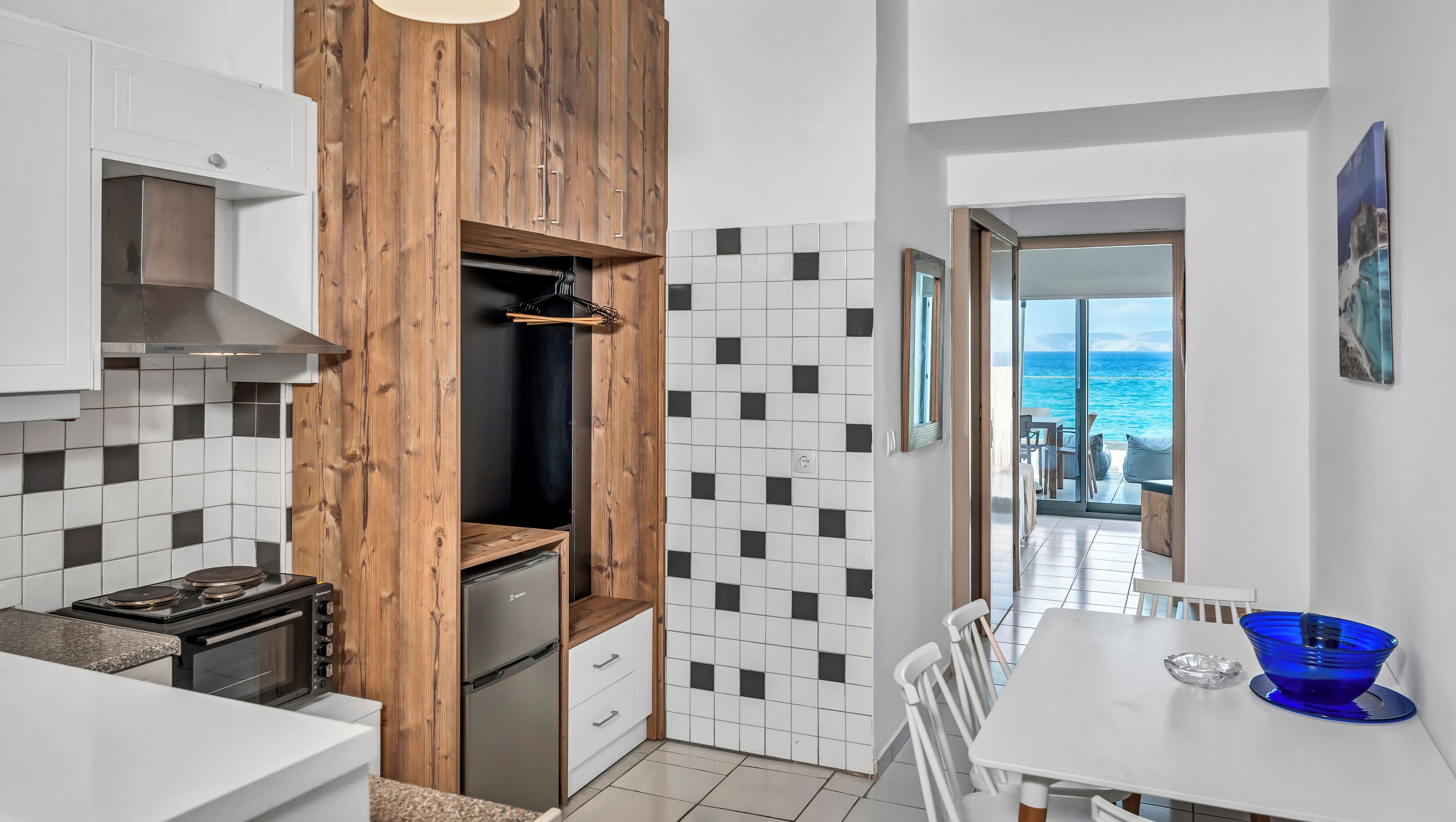 A modern kitchen with a wooden accent wall, a view of the sea through a sliding door, and a dining area with a blue bowl.