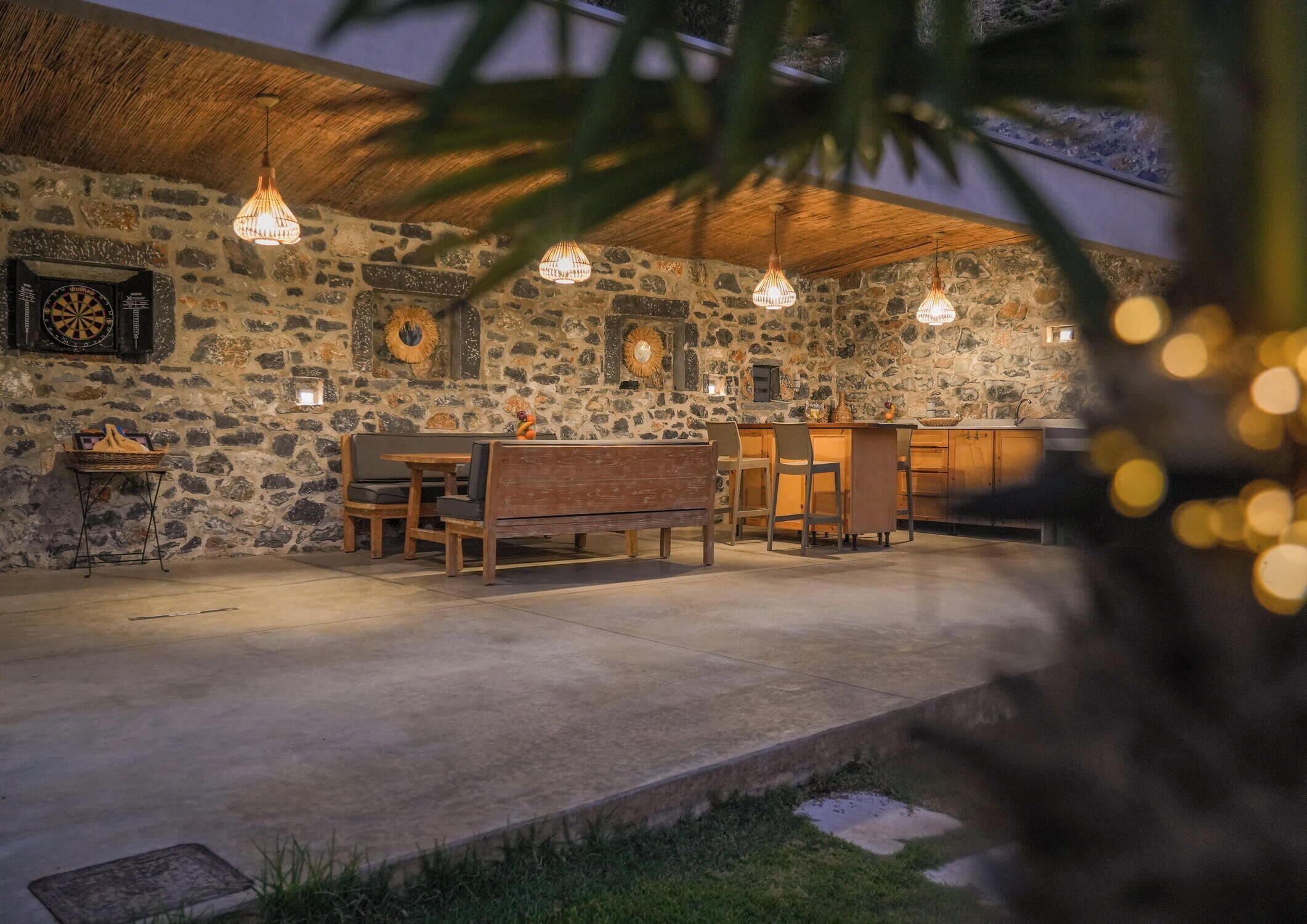 An outdoor dining area with stone walls, wooden furniture, and hanging pendant lights.