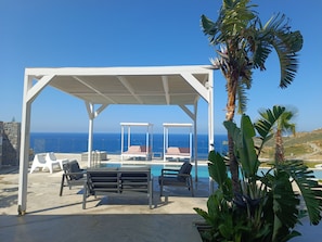 Un área de estar techada al aire libre con piscina, pérgola blanca y sillones de descanso.