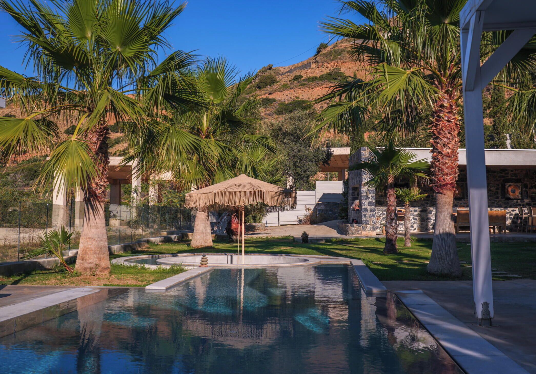 A swimming pool with a thatched umbrella, palm trees, and a stone building in the background.