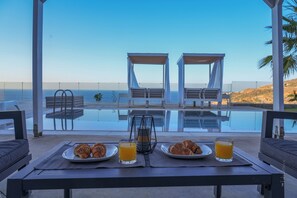 Une table à manger, dressée avec des assiettes de nourriture et des boissons, donnant sur une piscine et la mer.