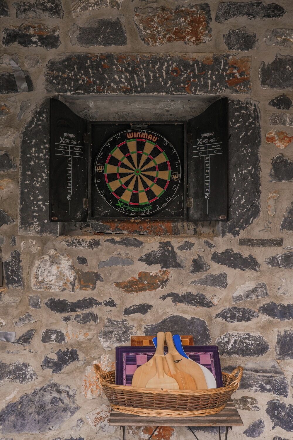 A dartboard mounted on a stone wall, a dartboard cabinet, and a basket with wooden paddles.