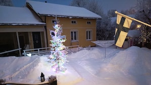 Paisaje nevado con un árbol de Navidad iluminado y una casa al fondo.