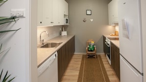 Cocina compacta con gabinetes blancos, microondas, pileta y lavavajillas. Hay una alfombra pequeña en el piso y un reloj decorativo en la pared.