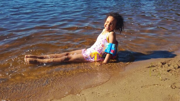 Un niño sentado en una playa arenosa cerca de una masa de agua, con un puente al fondo.