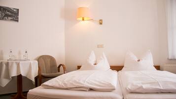 A hotel room with two beds, a table with glasses and a bottle, a chair, and a wall-mounted lamp.