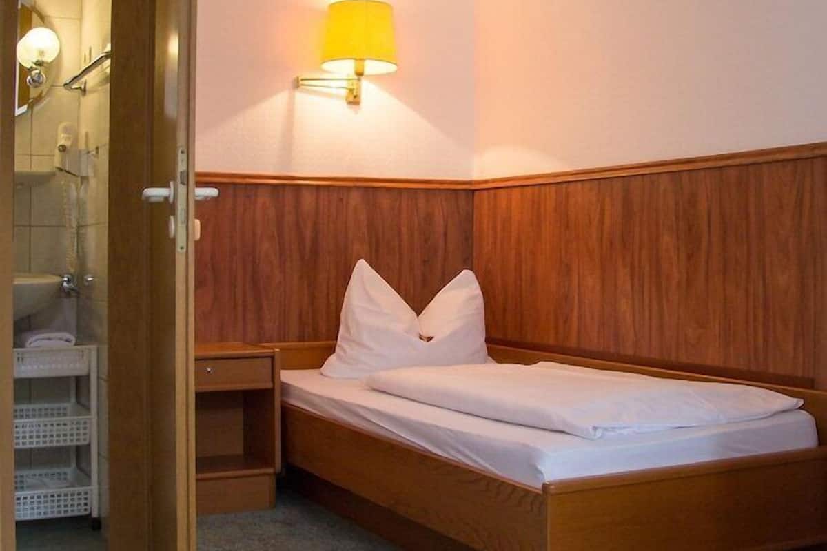 Un lit simple avec une tête de lit en bois, une table de chevet, une lampe fixée au mur et une salle de bain visible à travers une porte ouverte.