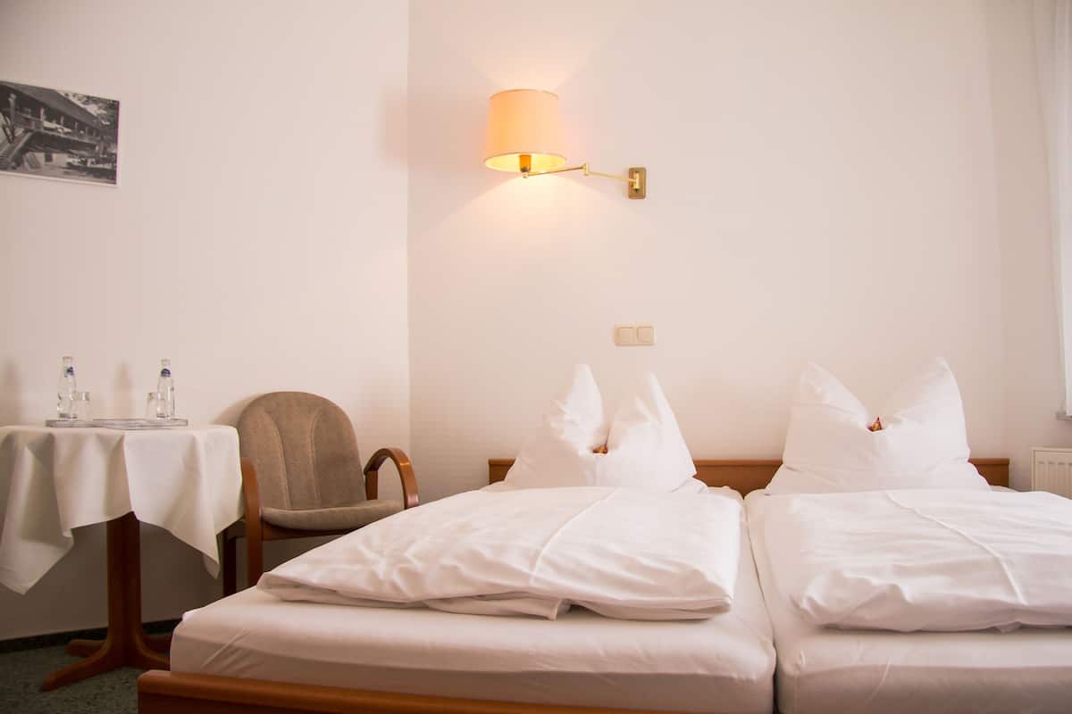 Une chambre d’hôtel avec deux lits, une chaise, une table recouverte d’une nappe blanche, une lampe fixée au mur et un tableau encadré accroché au mur.