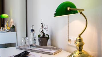 Ein Schreibtisch mit einer grünen Schreibtischlampe, einer Fernbedienung, einer Glasflasche, einem Topfpflanzen und einem gerahmten Bild an der Wand.