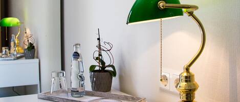 Un escritorio con una lámpara de escritorio verde, un control remoto, una botella de vidrio, una planta en maceta y un cuadro en la pared.