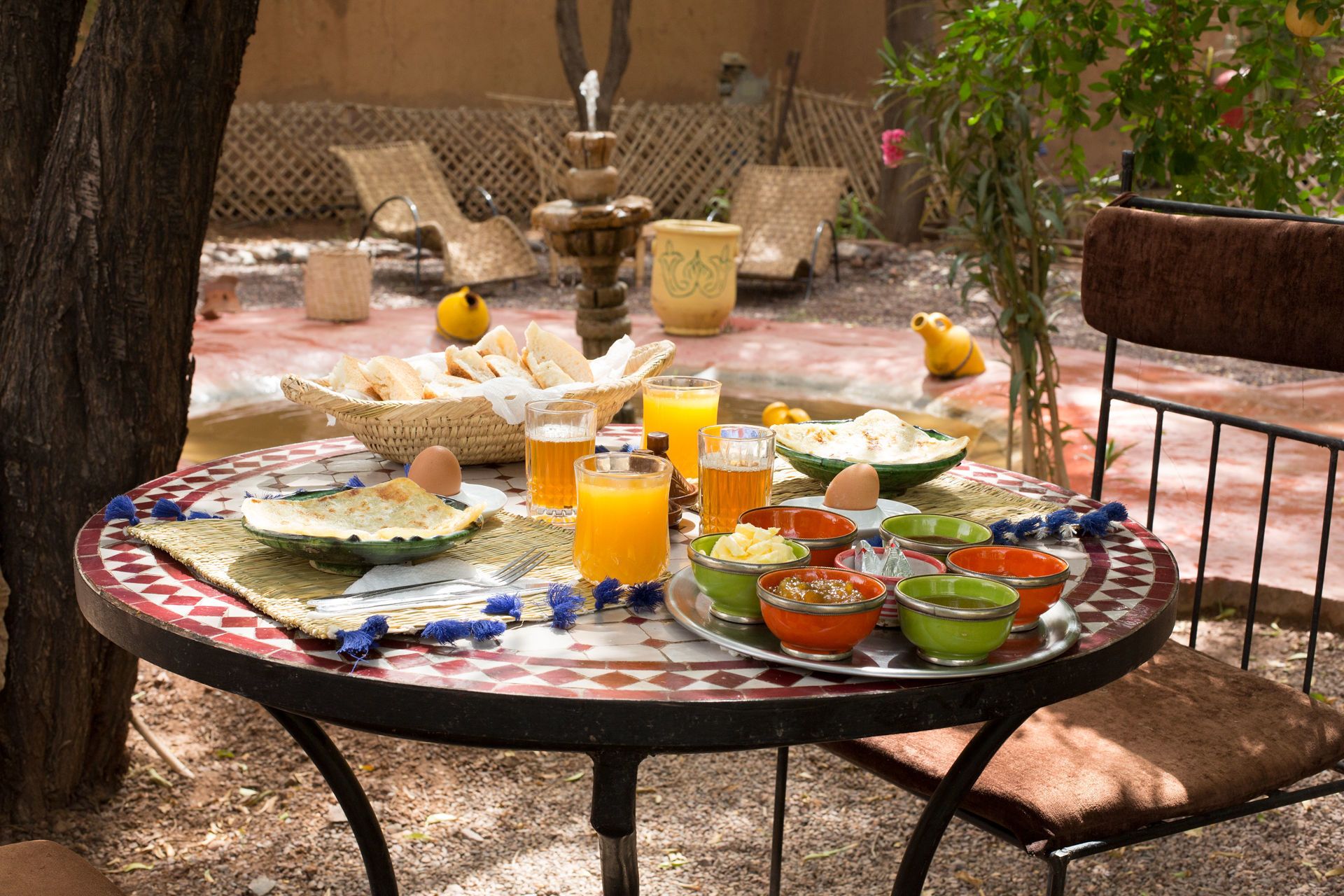 A round table with a checkered tablecloth, various dishes, glasses of orange juice and beer, and a basket of bread.