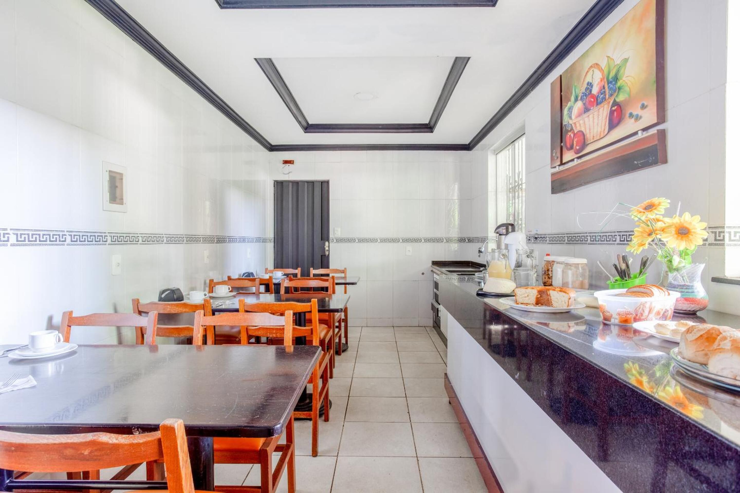 A dining area with wooden tables and chairs, a kitchen counter with food items, and a painting on the wall.