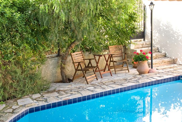 Un área de piscina con baldosas de piedra, sillas de madera y plantas en macetas.