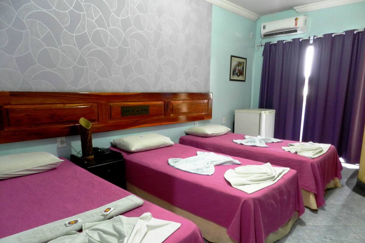 A hotel room with two beds, a wooden headboard, a small refrigerator, and a window with purple curtains.