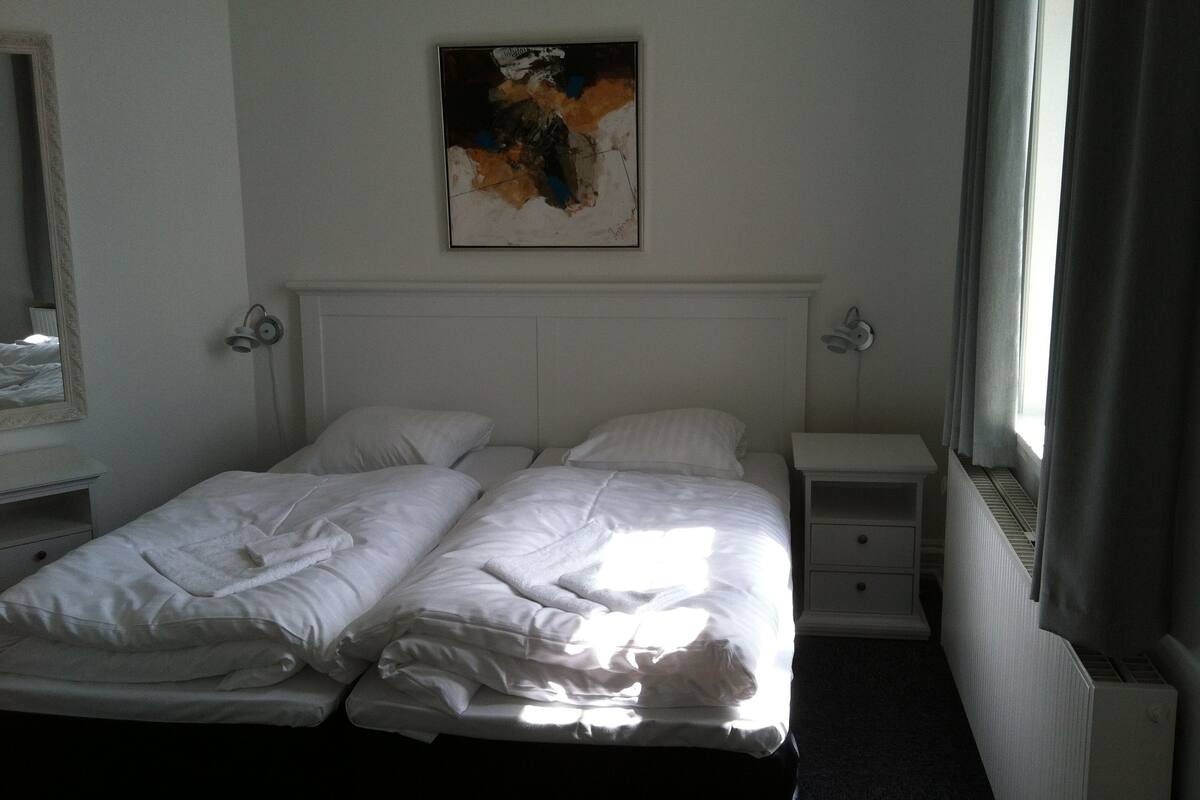 A hotel room with two single beds, a nightstand, a bedside table, a mirror, and a painting on the wall.