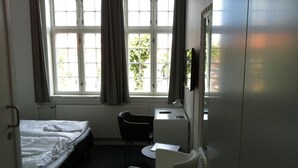 Quarto de hotel pequeno com duas camas de solteiro, uma escrivaninha com cadeira, um radiador e uma janela com cortinas.
