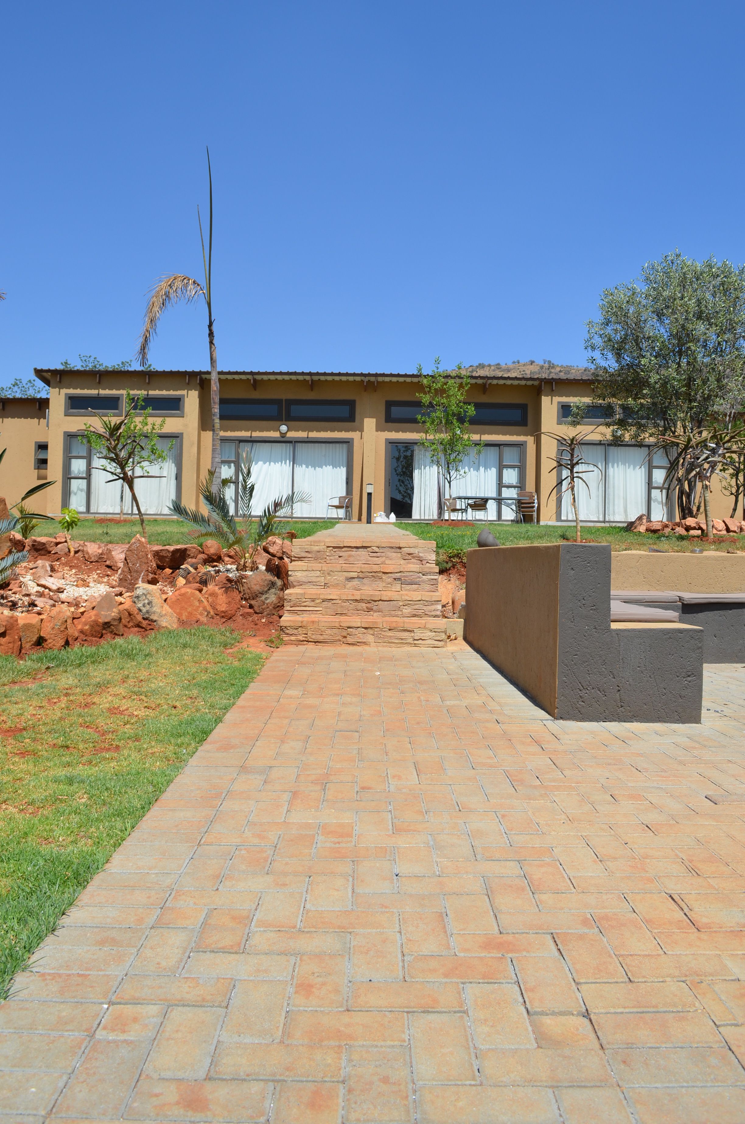 A pathway leading to a building with a modern design, featuring a stone wall and a small garden area with various plants.